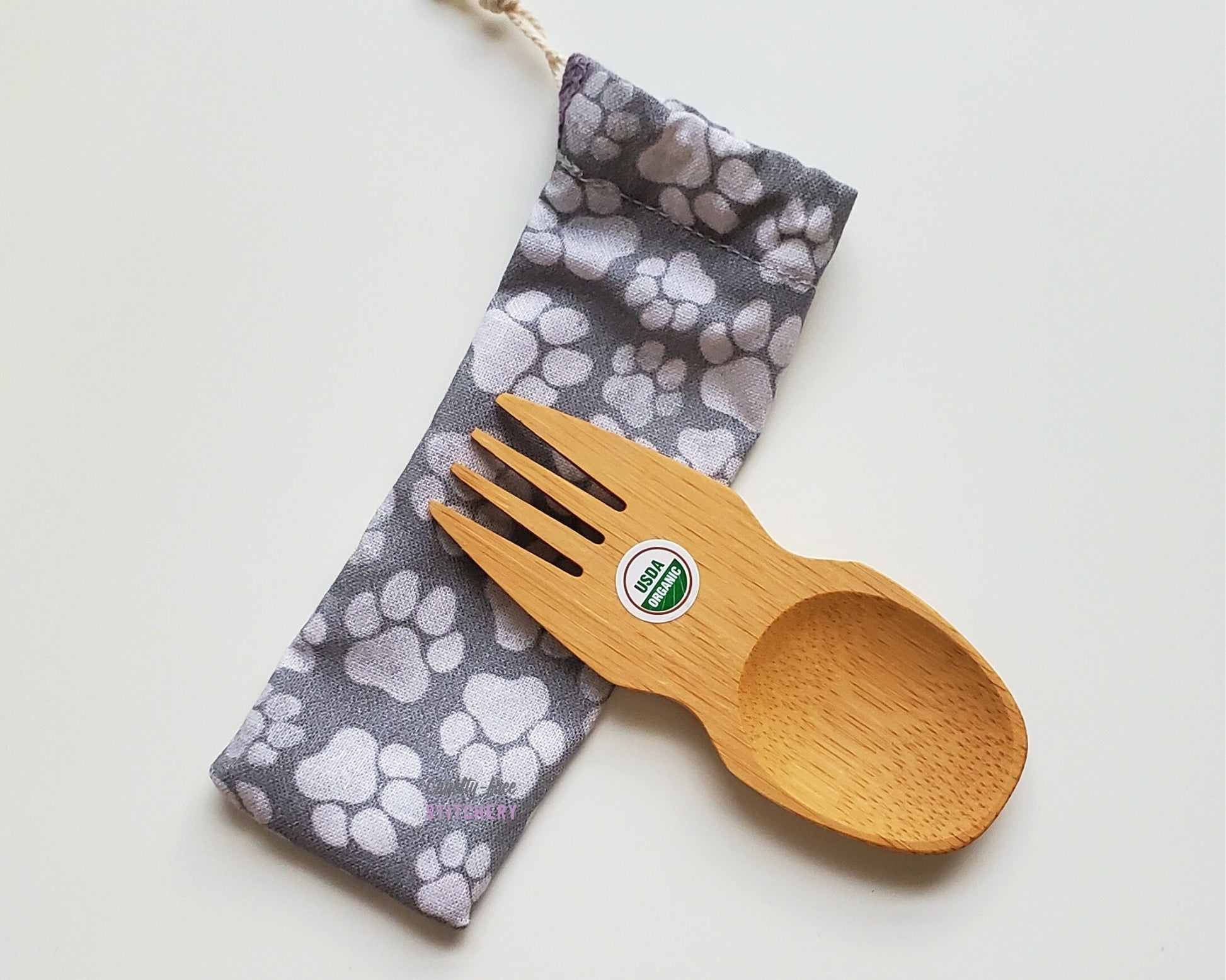 Reusable bamboo spork with pouch. The pouch is a grey fabric with various sizes of faded white paw prints. The pouch is sitting diagonally with the spork partially on top pointing the other way. The spork is small, a double ended fork and spoon.