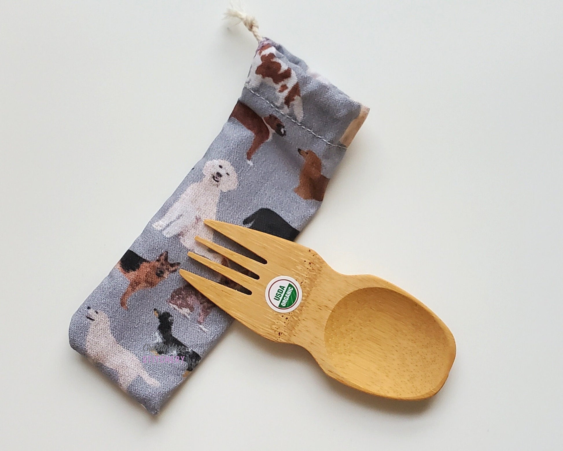 Reusable bamboo spork with pouch. The pouch is grey with various dogs printed on. The pouch is sitting diagonally with the spork partially on top pointing the other way. The spork is small, a double ended fork and spoon.