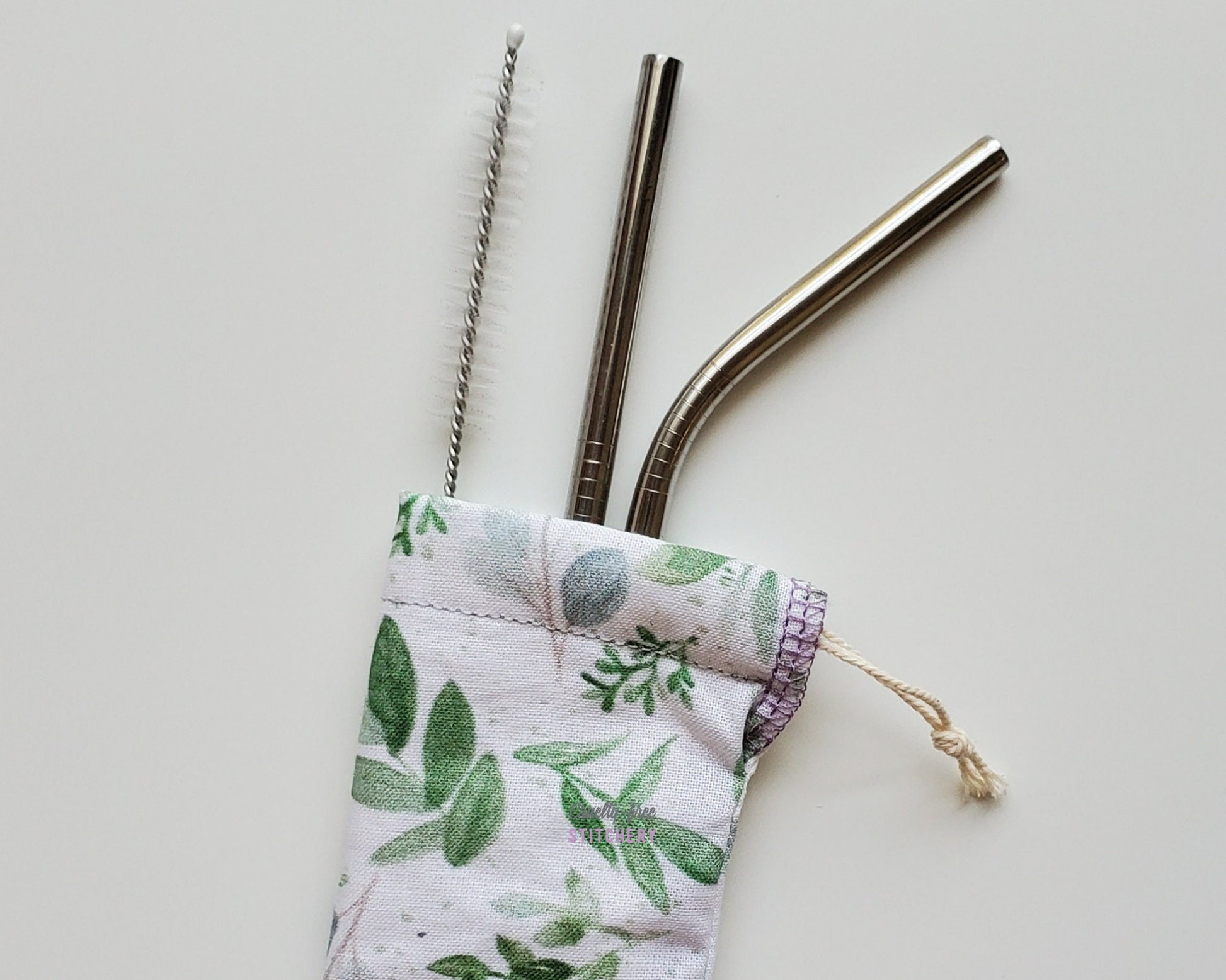 Reusable straw pouch with stainless steel straws sticking out the top. The fabric of the pouch is white with green eucalyptus vines and leaves printed on, and it has a drawstring closure.