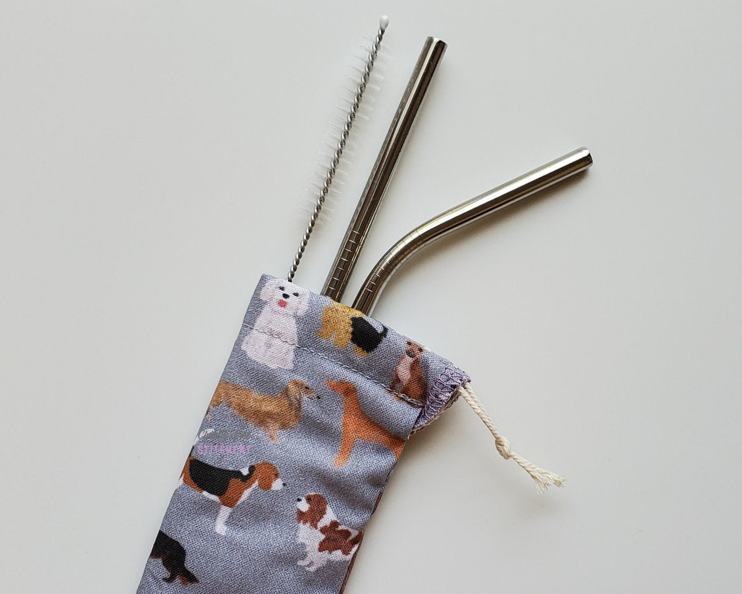 Reusable straw pouch with stainless steel straws sticking out the top. The fabric of the pouch is grey with various dogs printed on, such as beagles, basset hounds, yorkies, etc, and it has a drawstring closure.