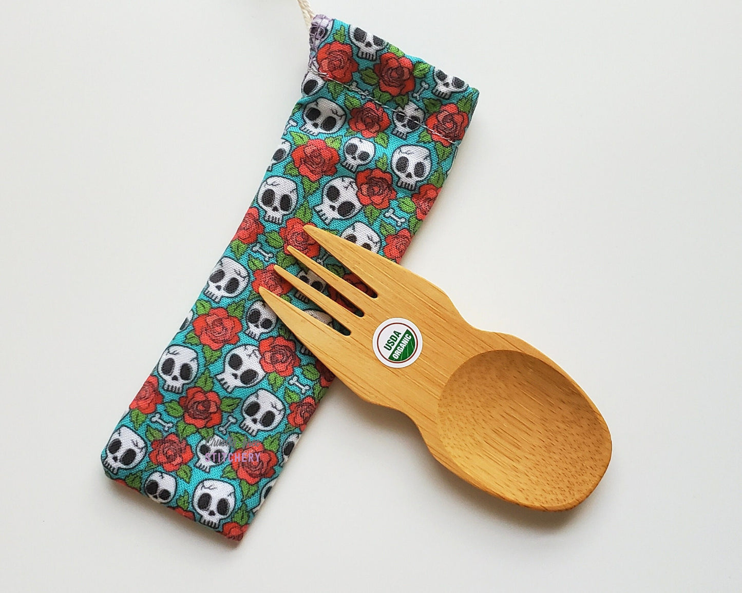 Reusable bamboo spork with pouch. The pouch is a blue fabric with tiny white skulls and red roses. The pouch is sitting diagonally with the spork partially on top pointing the other way. The spork is small, a double ended fork and spoon.