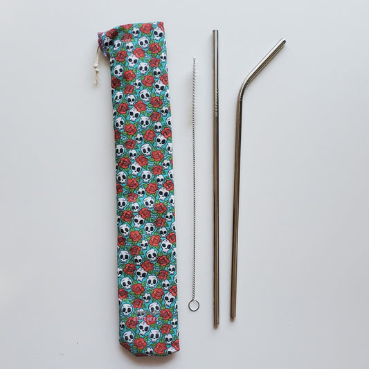 Reusable straw pouch in the same skull and roses print laying vertically next to a straw cleaner brush, a straight stainless steel straw, and a bent stainless steel straw.
