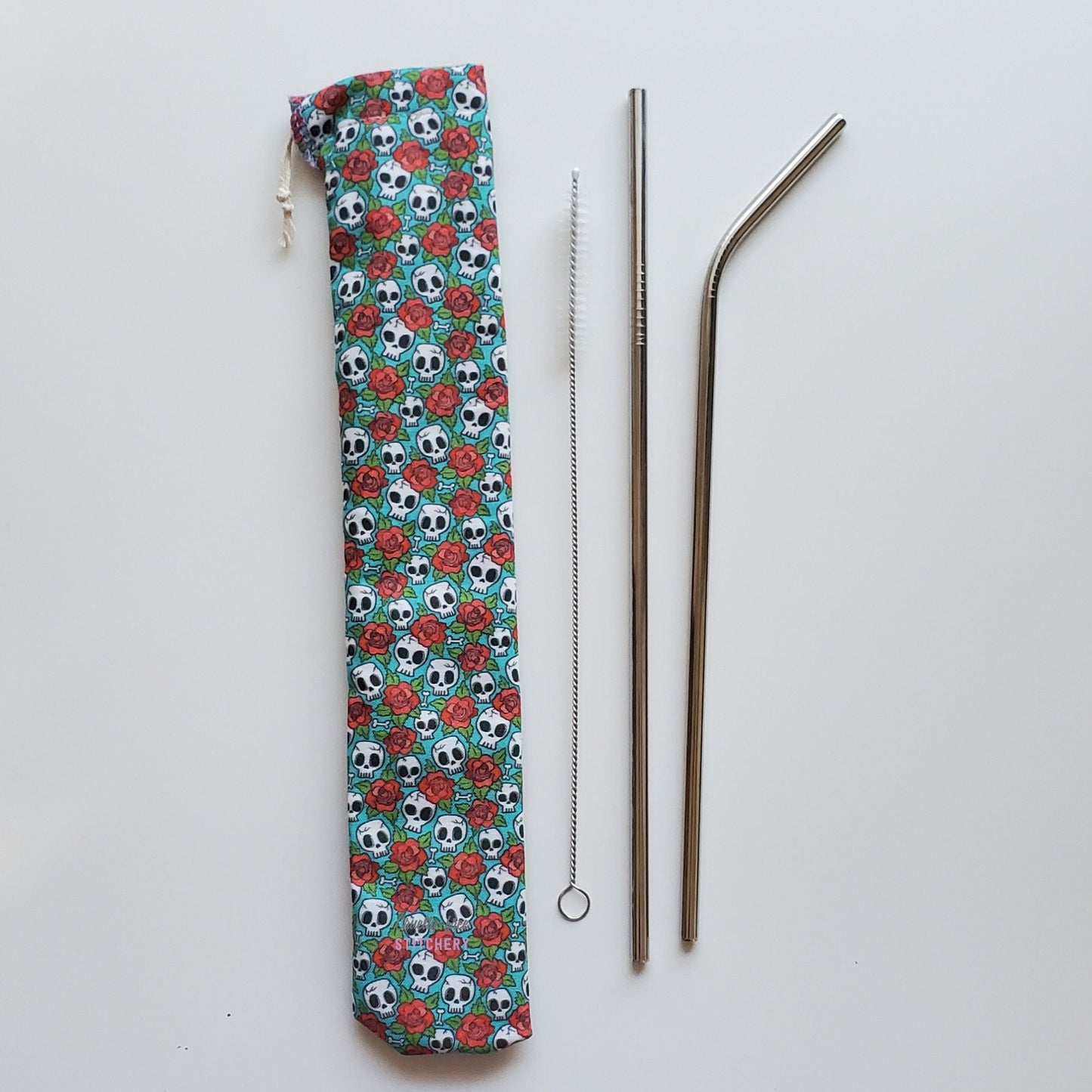 Reusable straw pouch in the same skull and roses print laying vertically next to a straw cleaner brush, a straight stainless steel straw, and a bent stainless steel straw.