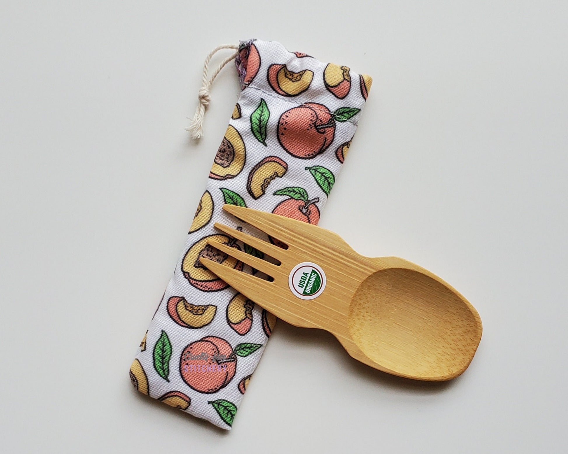 Reusable bamboo spork with pouch. The pouch is a white fabric with small peaches, some whole and some slices. The pouch is sitting diagonally with the spork partially on top pointing the other way. The spork is small, a double ended fork and spoon.
