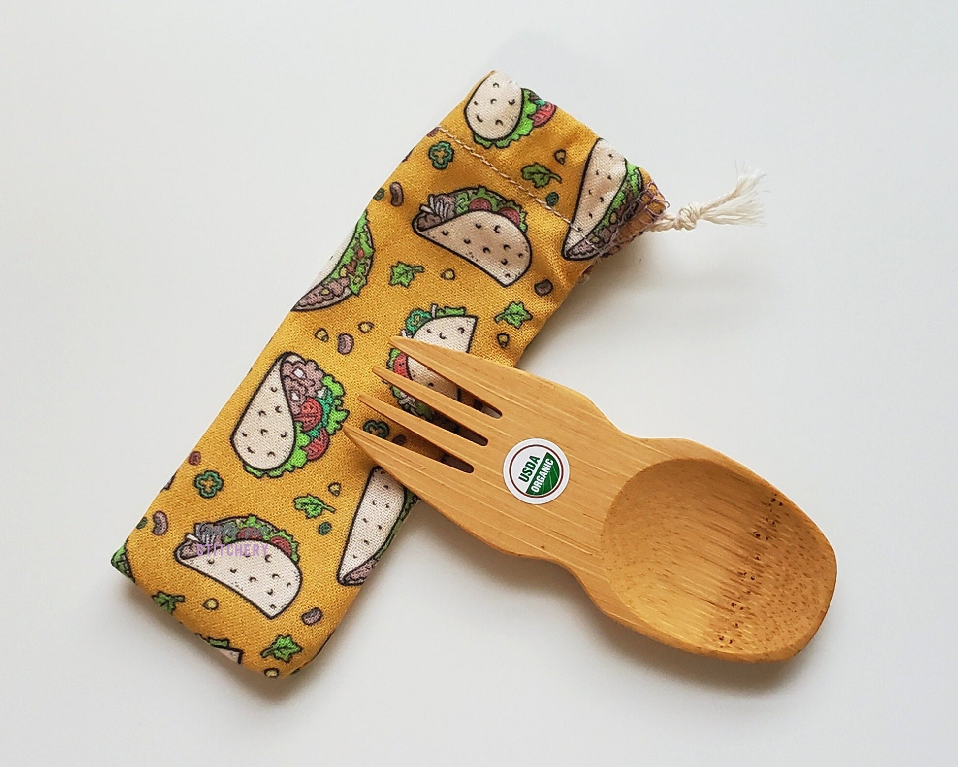 Reusable bamboo spork with pouch. The pouch is a mustard yellow fabric with small tacos printed on. The pouch is sitting diagonally with the spork partially on top pointing the other way. The spork is small, a double ended fork and spoon.