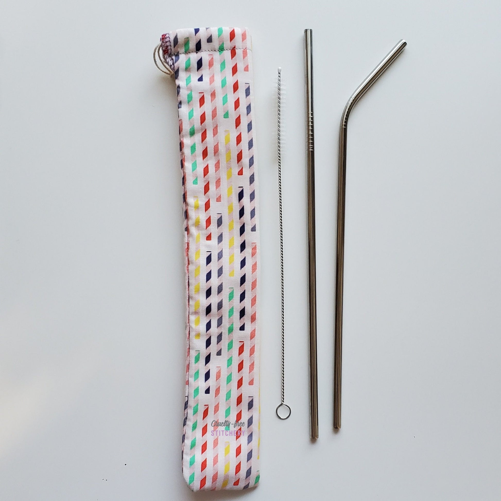 Reusable straw pouch in the same paper straw print laying vertically next to a straw cleaner brush, a straight stainless steel straw, and a bent stainless steel straw.
