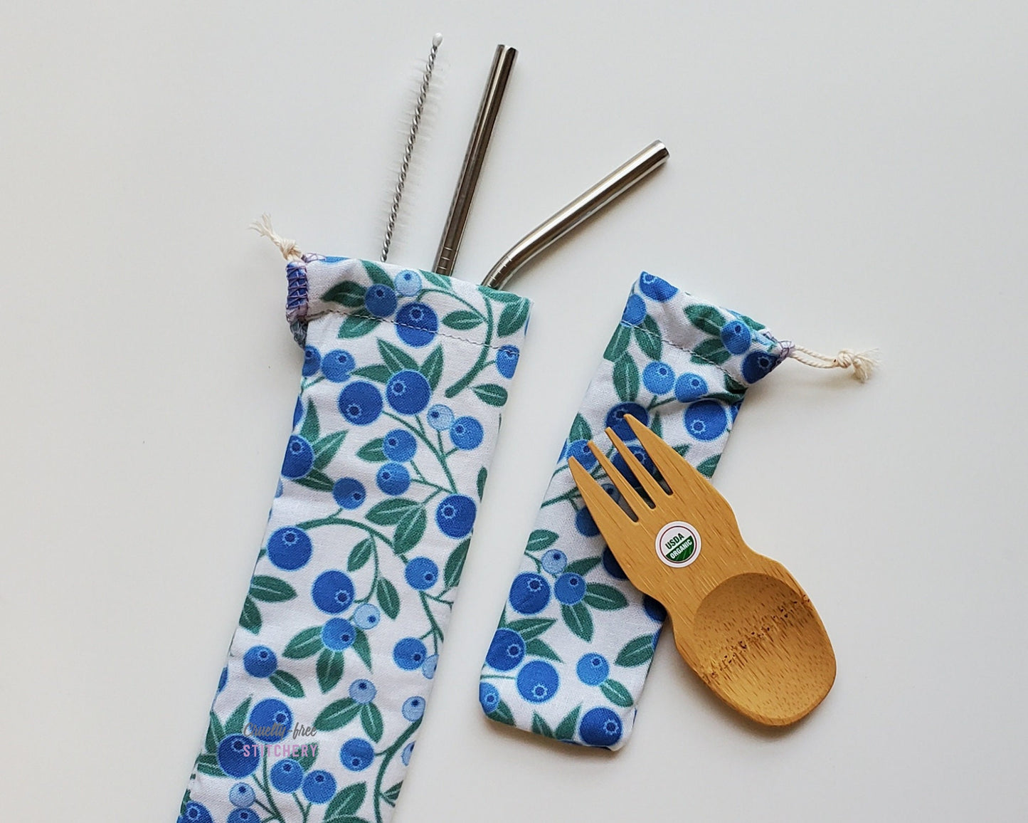 Reusable bamboo spork and straw pouch set. The pouches are a white fabric with printed blueberries and green vines and leaves.