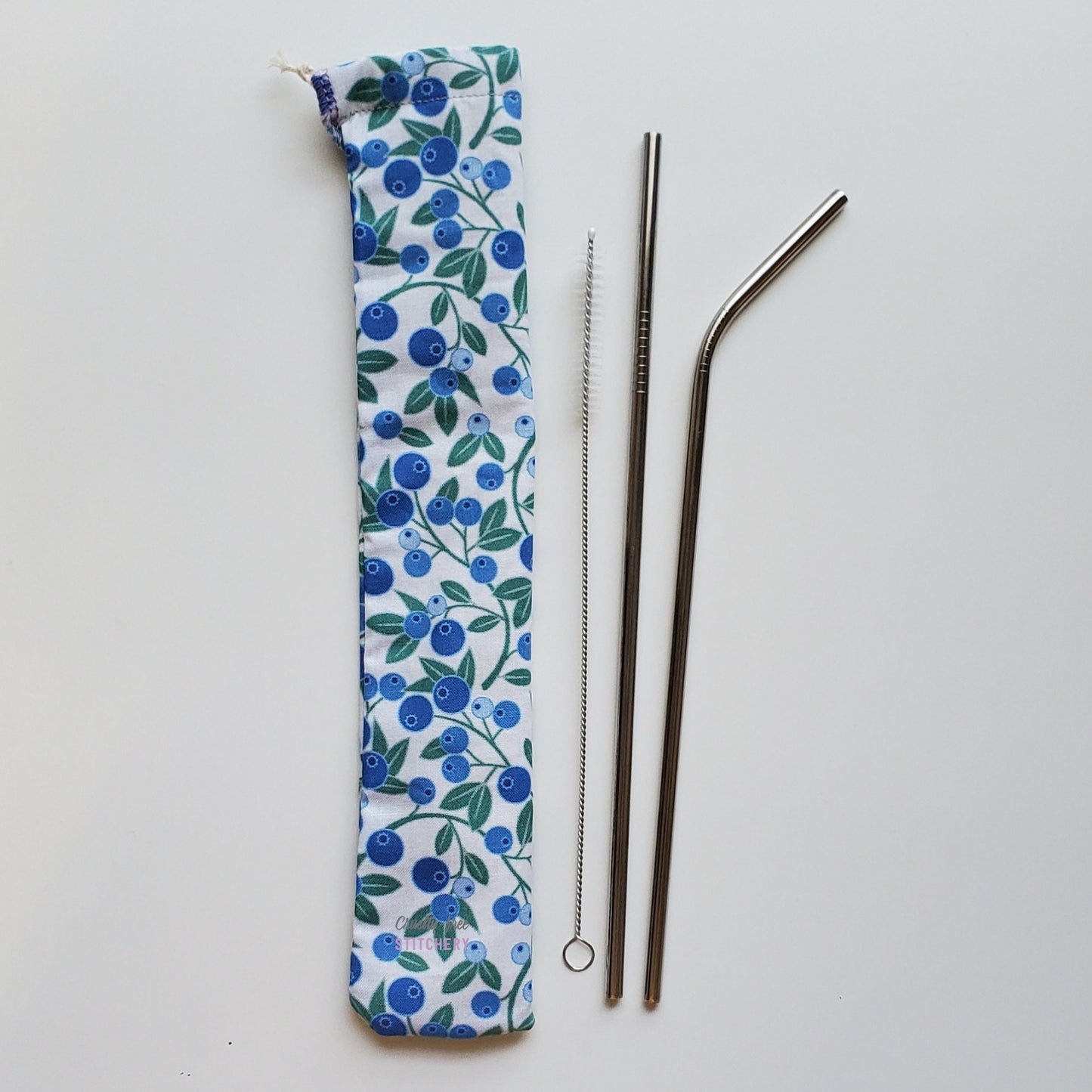 Reusable straw pouch in the same blueberry print laying vertically next to a straw cleaner brush, a straight stainless steel straw, and a bent stainless steel straw.