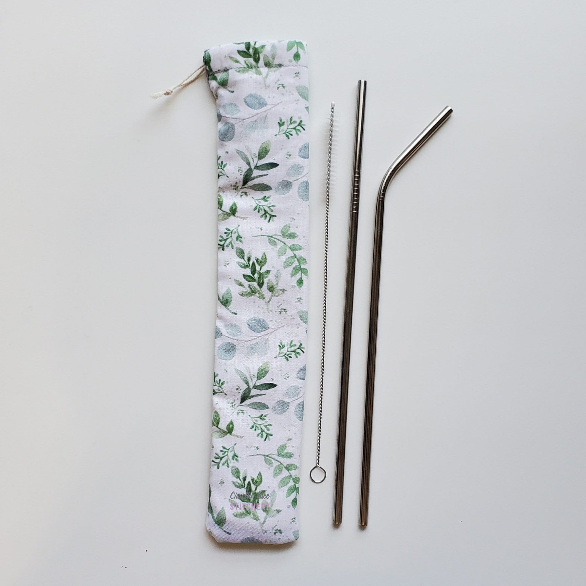 Reusable straw pouch in the same white with eucalyptus leaves print laying vertically next to a straw cleaner brush, a straight stainless steel straw, and a bent stainless steel straw.