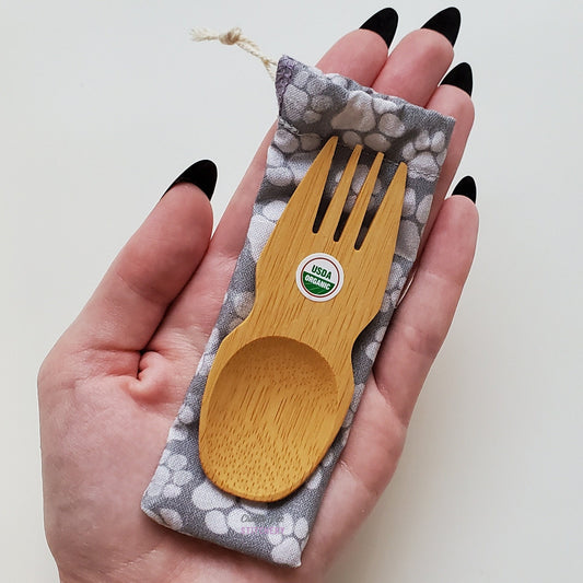Reusable bamboo spork with pouch. Paw print fabric pouch with bamboo spork on top, laid on a hand for size reference. The spork is slightly longer than the fingers.