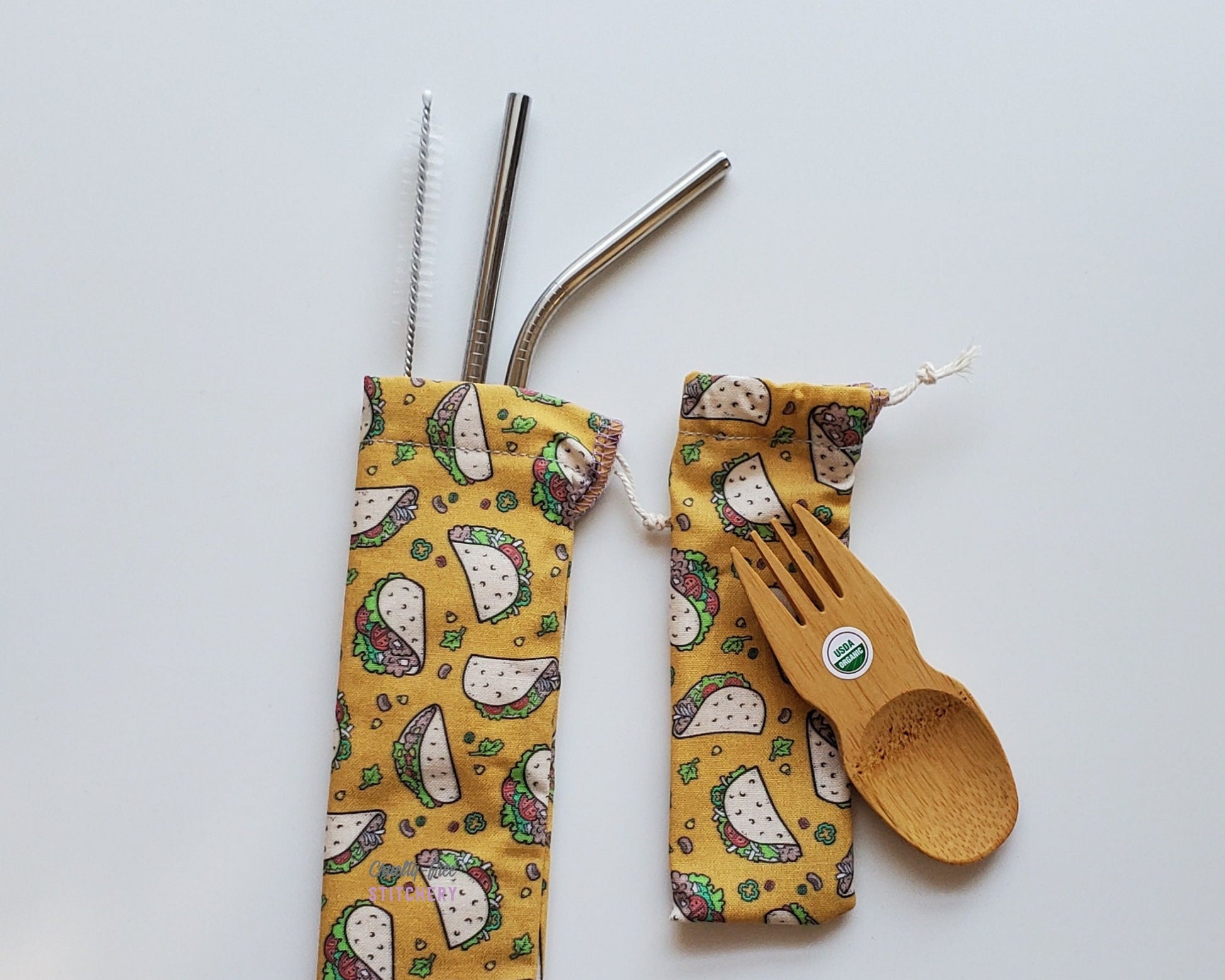 Reusable bamboo spork and stainless steel straw pouch set. The pouches are both a mustard yellow color with small cartoon tacos and fillings scattered around.