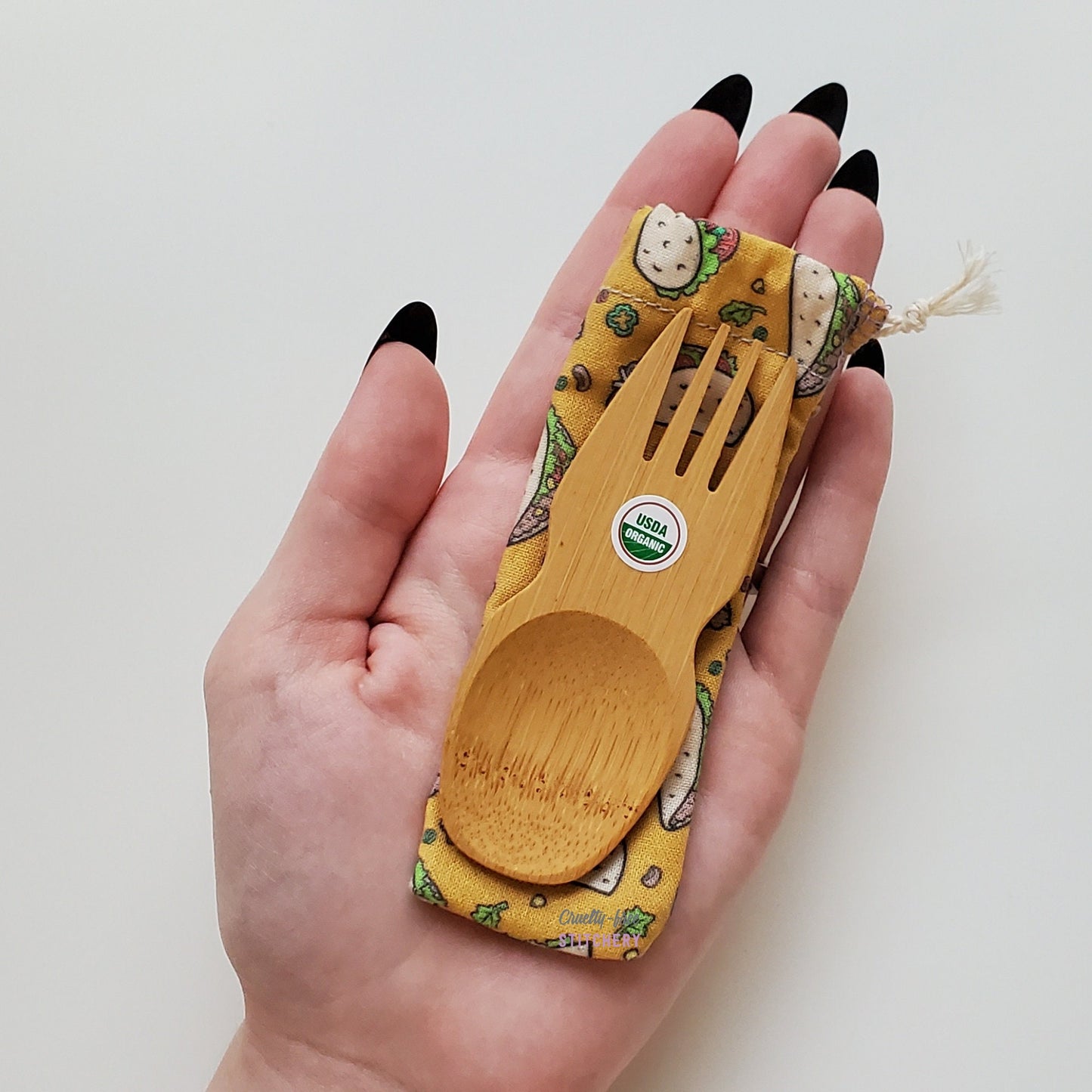 Reusable bamboo spork with pouch. Taco print fabric pouch with bamboo spork on top, laid on a hand for size reference. The spork is slightly longer than the fingers.