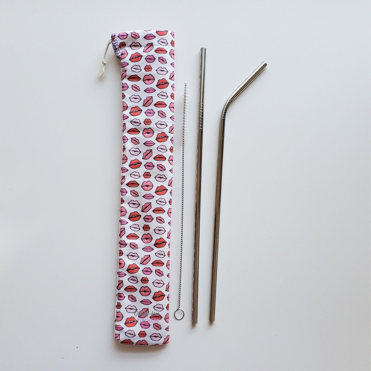 Reusable straw pouch in the same lips print laying vertically next to a straw cleaner brush, a straight stainless steel straw, and a bent stainless steel straw.