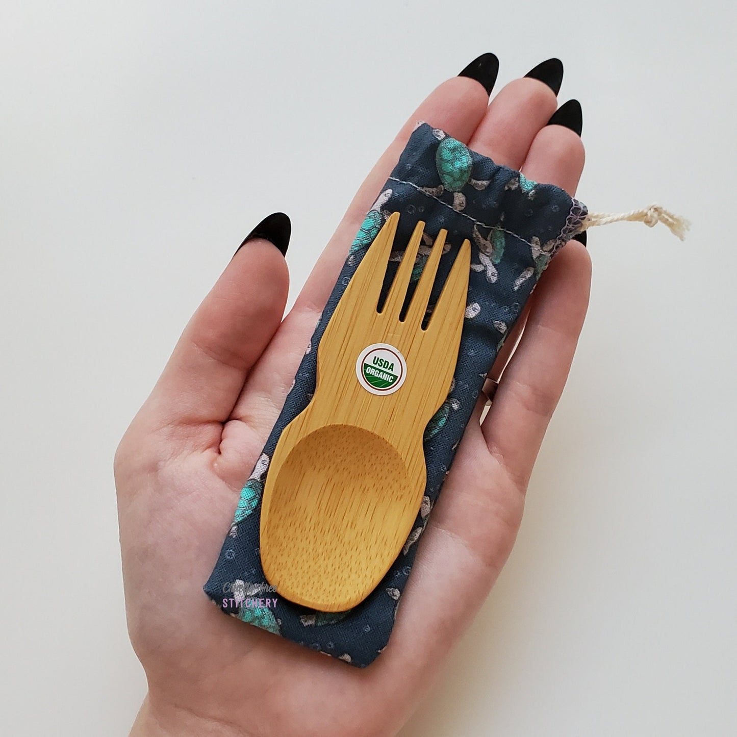 Reusable bamboo spork with pouch. Turtle print fabric pouch with bamboo spork on top, laid on a hand for size reference. The spork is slightly longer than the fingers.