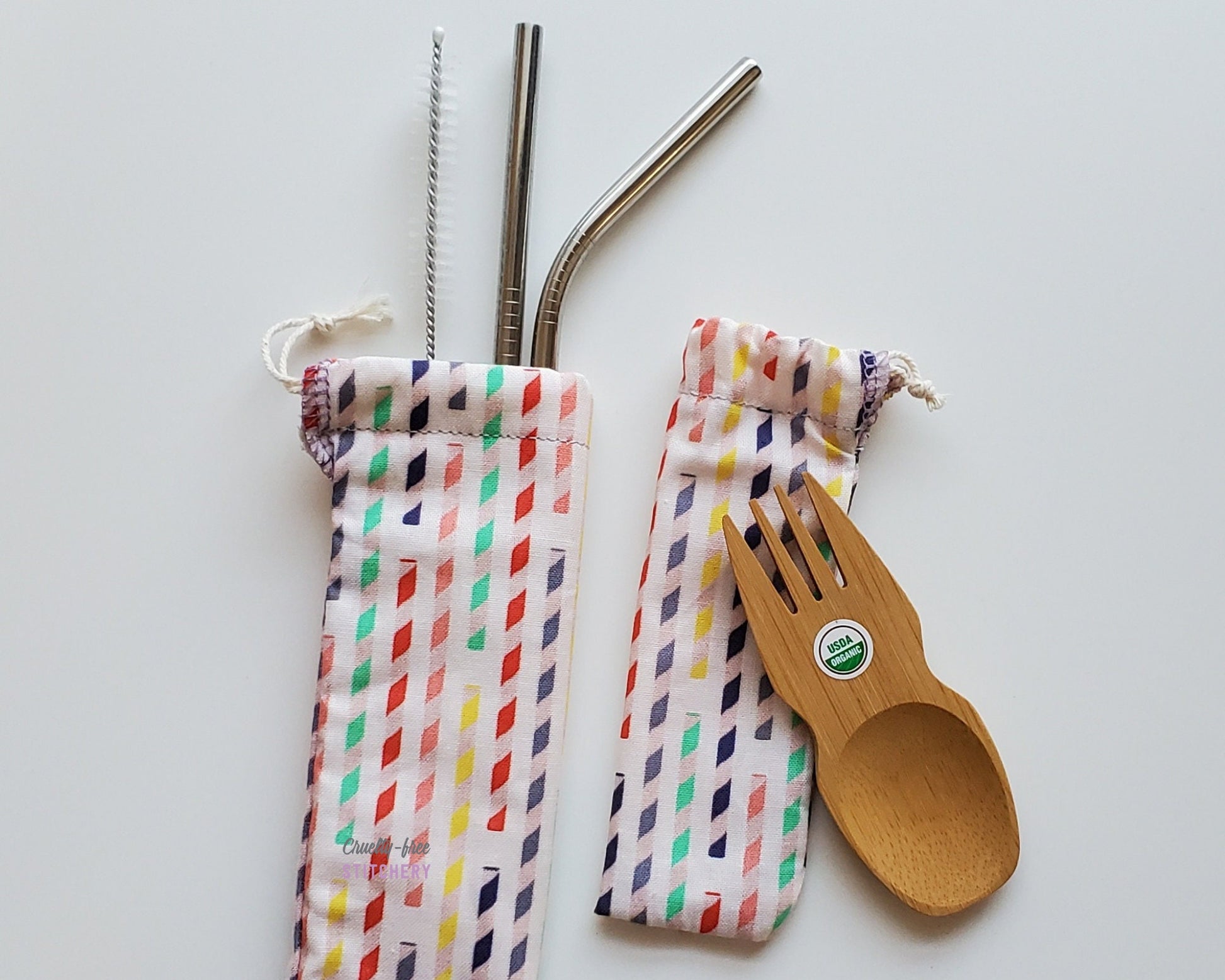Reusable bamboo spork and stainless steel straw pouch set. The pouches are both white with small striped paper straws printed on them. The paper straws are swirled with white and various colors like blue, red, pink, and yellow.