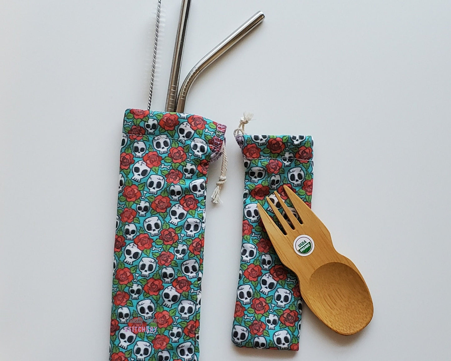 Reusable bamboo spork and stainless steel straw pouch set. The pouches are both teal blue with tiny cartoon skulls and red roses printed all over.