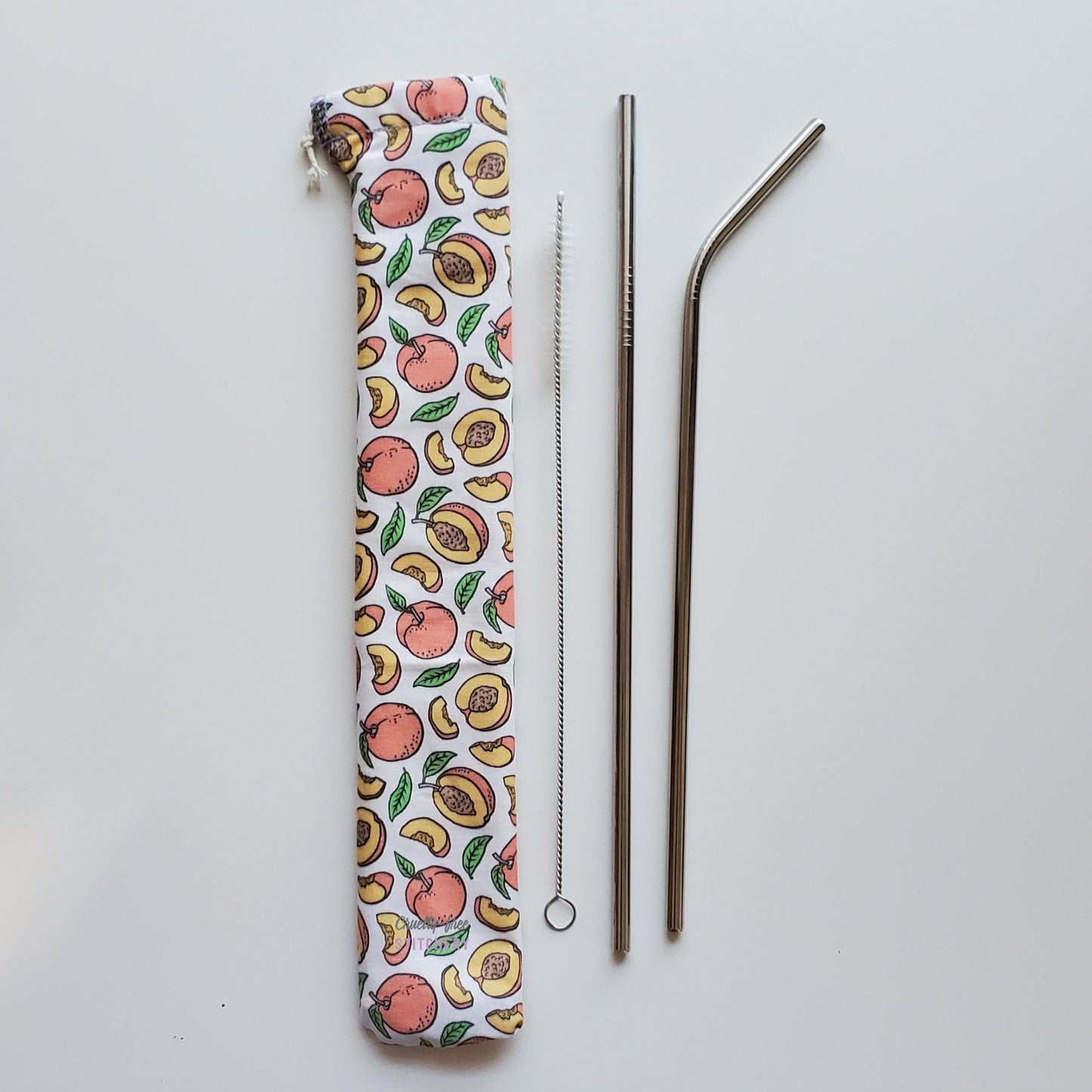 Reusable straw pouch in the same peaches print laying vertically next to a straw cleaner brush, a straight stainless steel straw, and a bent stainless steel straw.