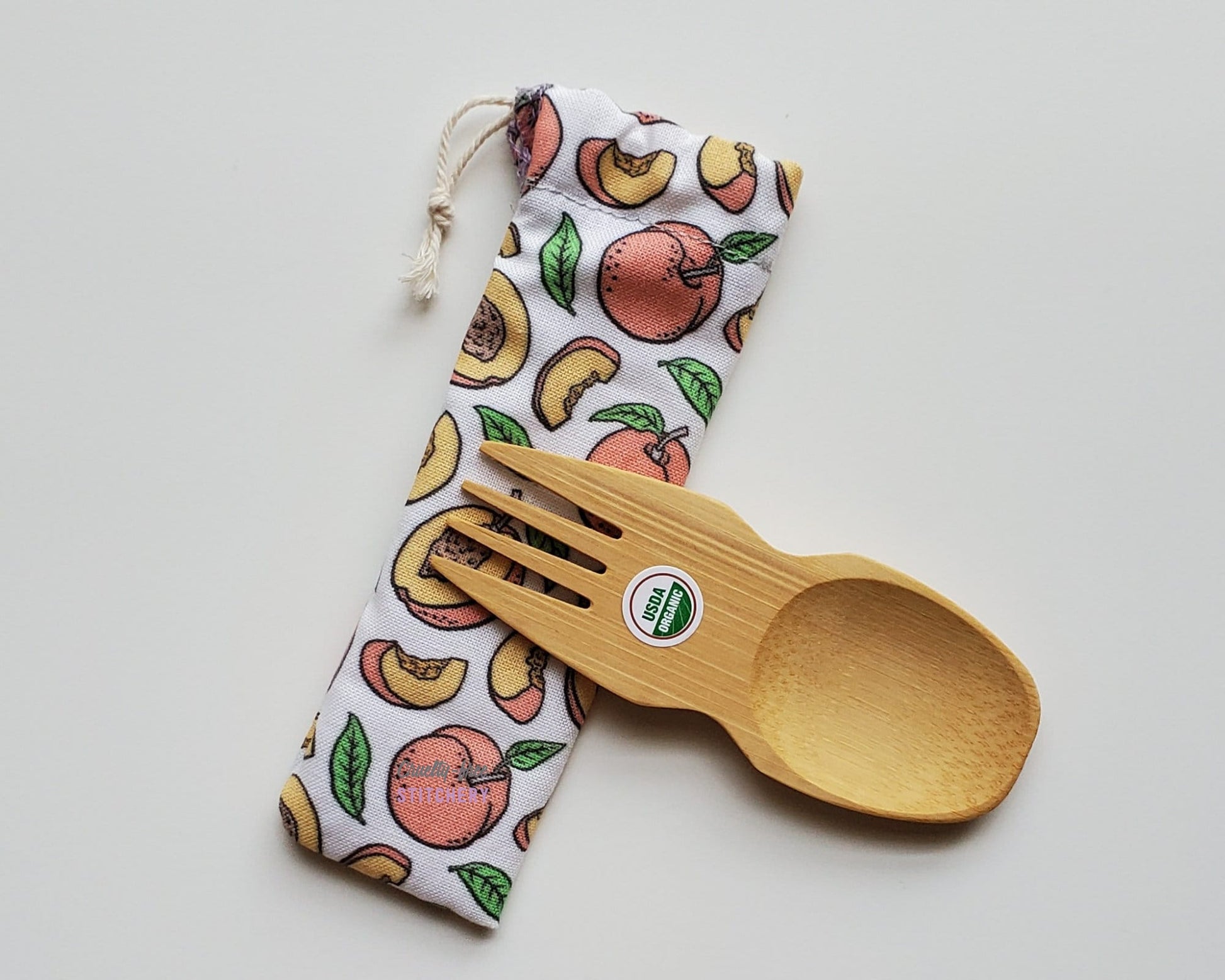 Peaches print reusable spork pouch. The pouch is sitting diagonally with the spork partially on top pointing the other way. The fork end of the spork is pointing to the left.