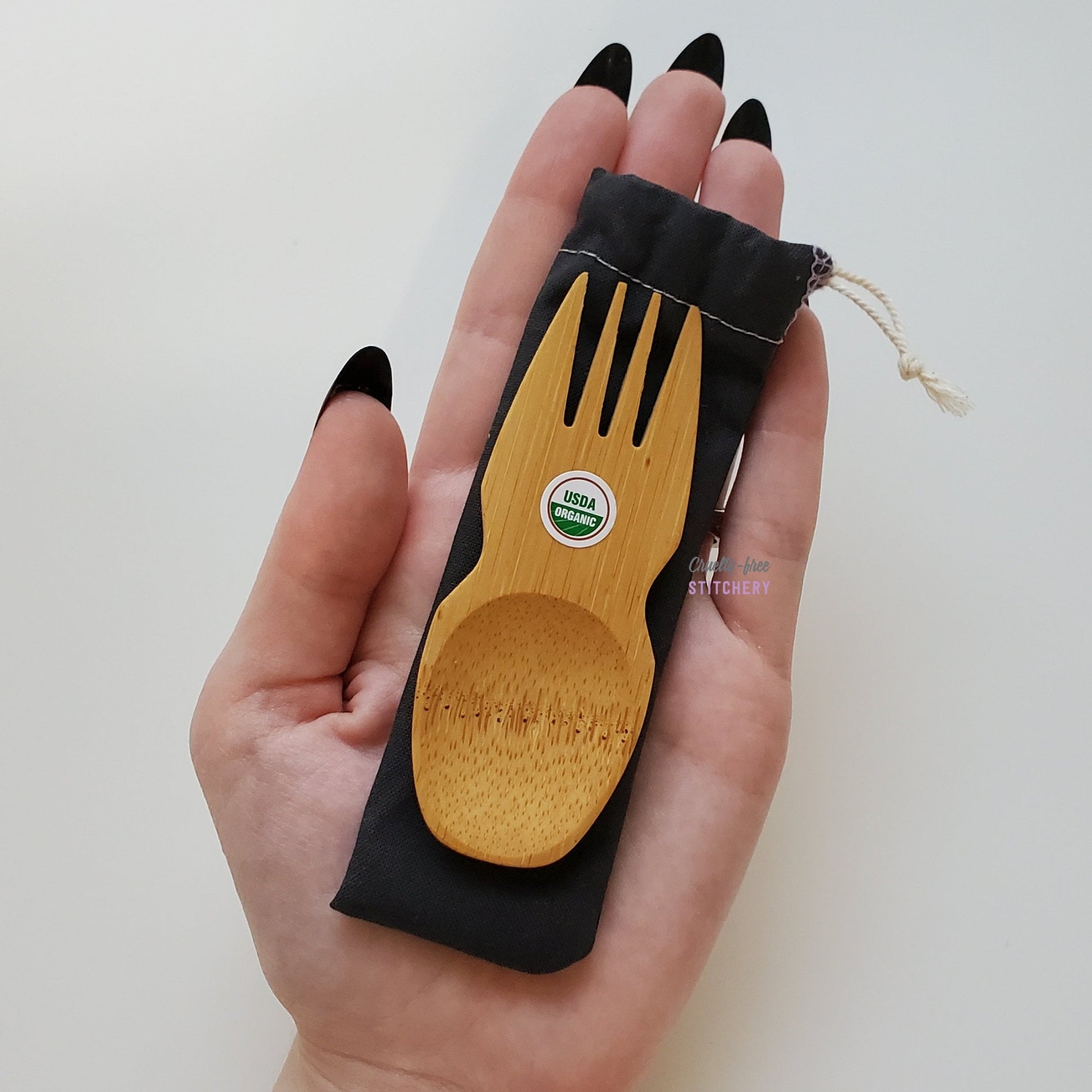 Reusable bamboo spork with pouch. Solid black fabric pouch with bamboo spork on top, laid on a hand for size reference. The spork is slightly longer than the fingers.