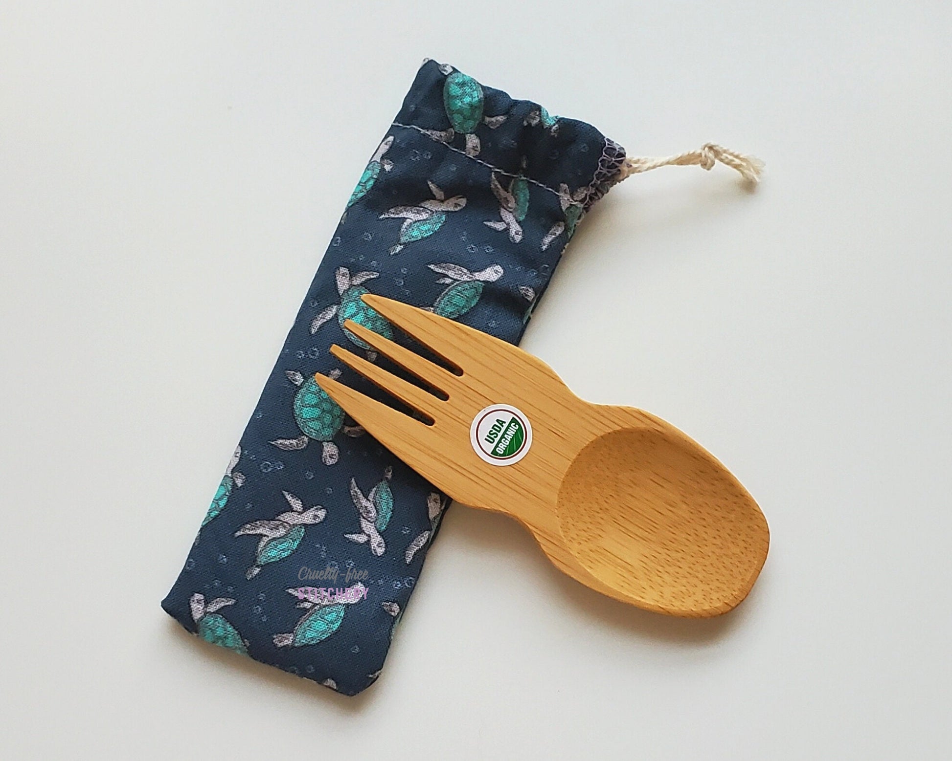 Reusable bamboo spork with pouch. The pouch is a dark navy blue fabric with tiny sea turtles. The pouch is sitting diagonally with the spork partially on top pointing the other way. The spork is small, a double ended fork and spoon.