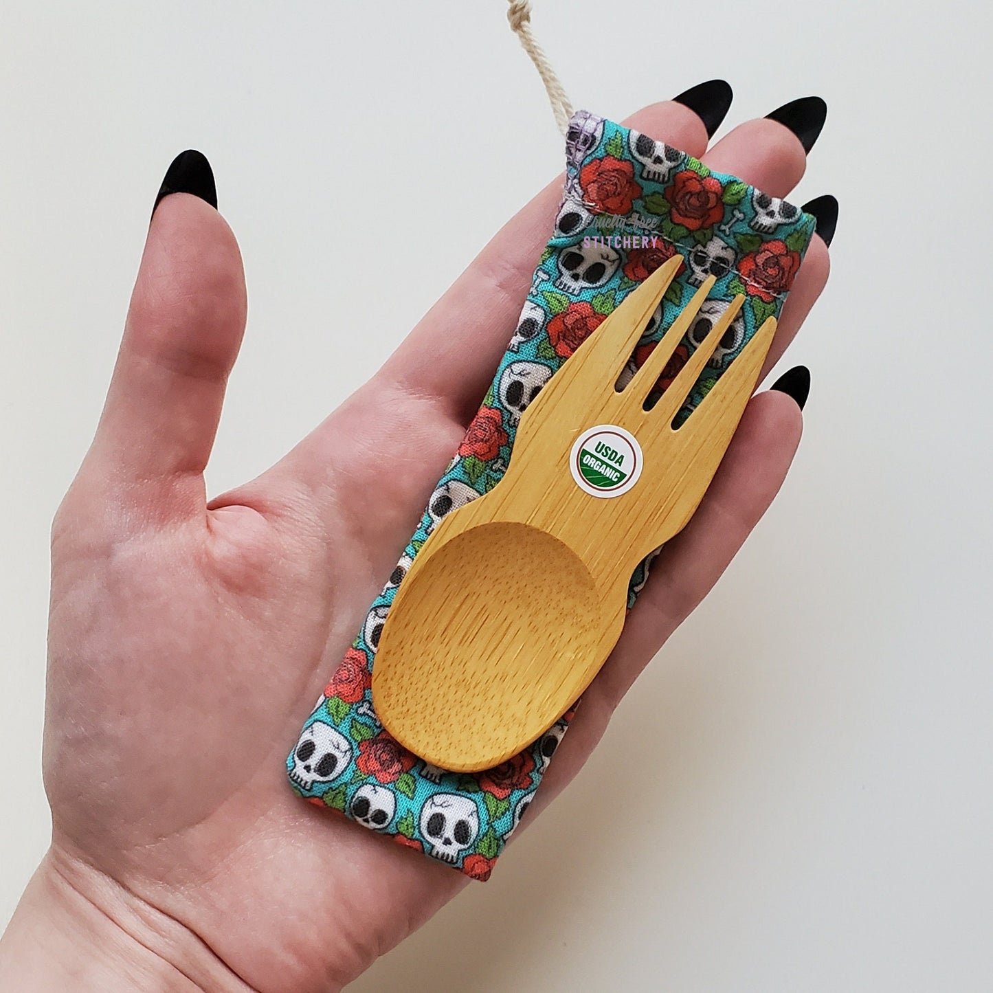 Reusable bamboo spork with pouch. Skull and roses print fabric pouch with bamboo spork on top, laid on a hand for size reference. The spork is slightly longer than the fingers.