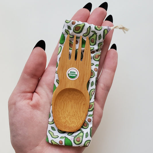 Reusable bamboo spork with pouch. Avocado print fabric pouch with bamboo spork on top, laid on a hand for size reference. The spork is slightly longer than the fingers.