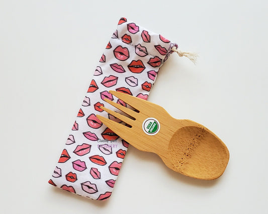 Reusable bamboo spork with pouch. The pouch is a white fabric with tiny lips in different shades of pink. The pouch is sitting diagonally with the spork partially on top pointing the other way. The spork is small, a double ended fork and spoon.