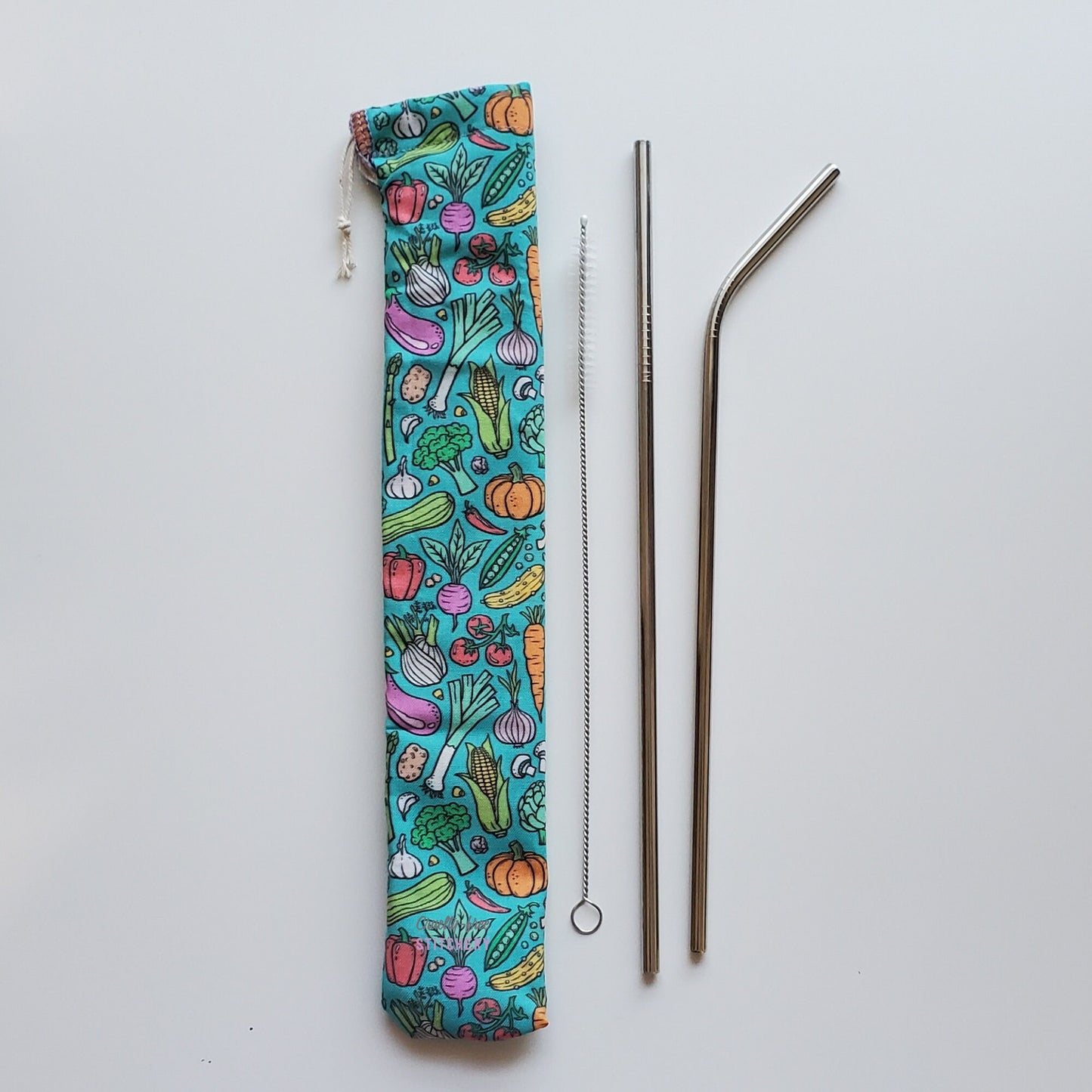 Reusable straw pouch in the same vegetable print laying vertically next to a straw cleaner brush, a straight stainless steel straw, and a bent stainless steel straw.