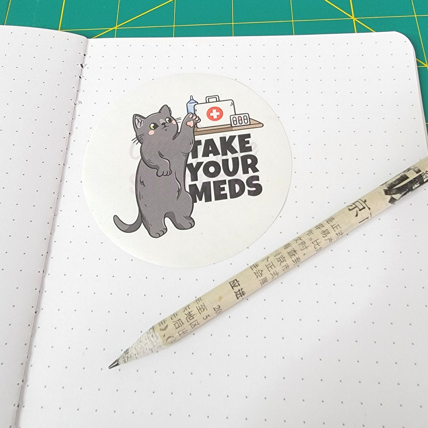 The &quot;take your meds&quot; sticker on a dot grid notebook next to a recycled newspaper pencil for size reference.