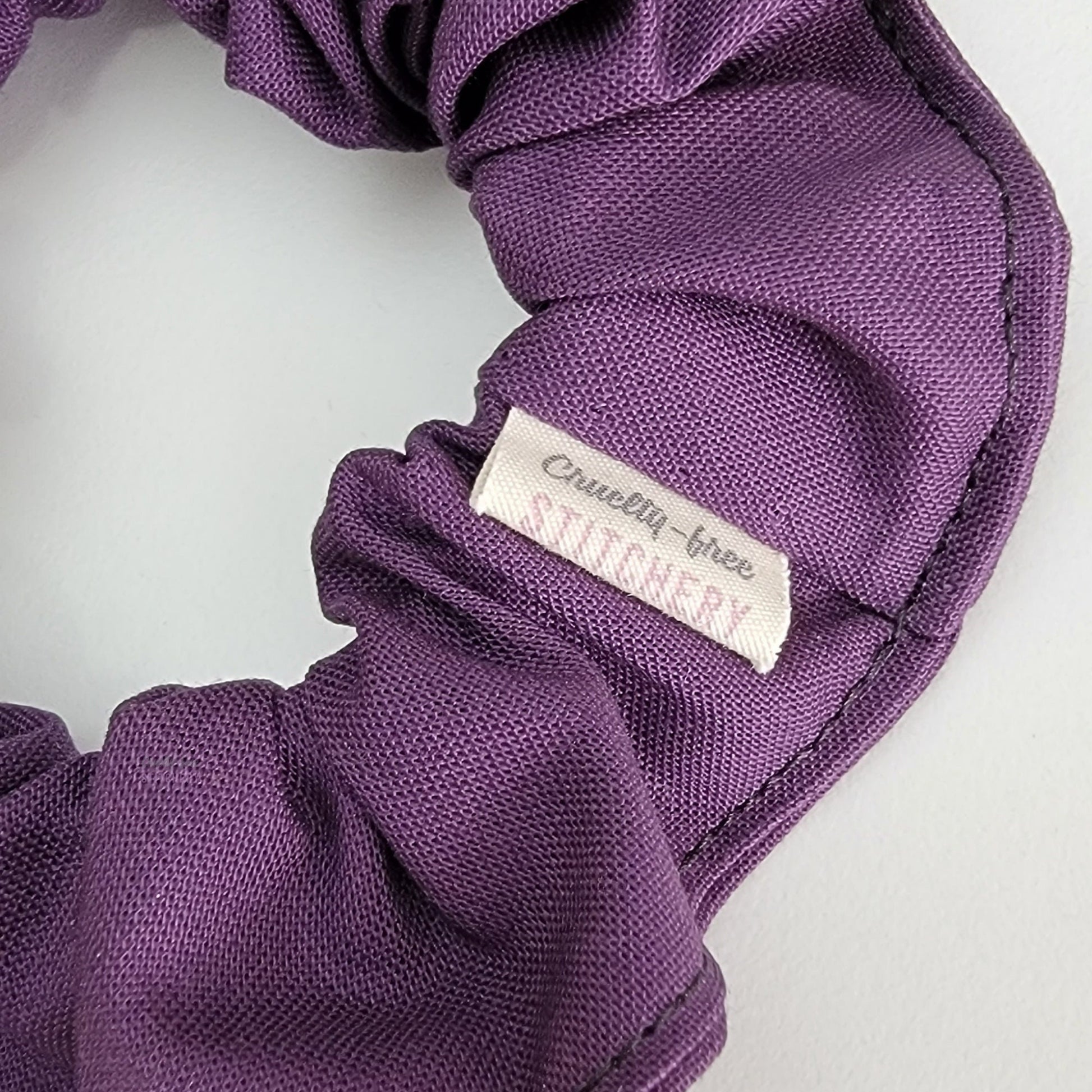Close-up of the purple scrunchie with a small white tag with the Cruelty-Free Stitchery logo.
