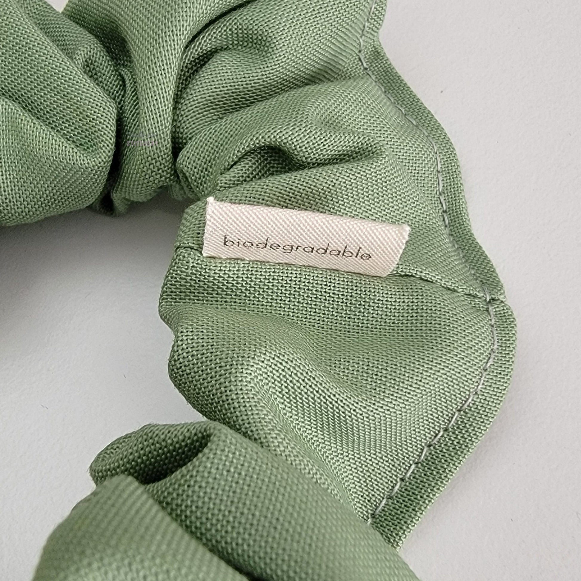 Close-up of a sage green scrunchie with the back of a small white tag that says "biodegradable"