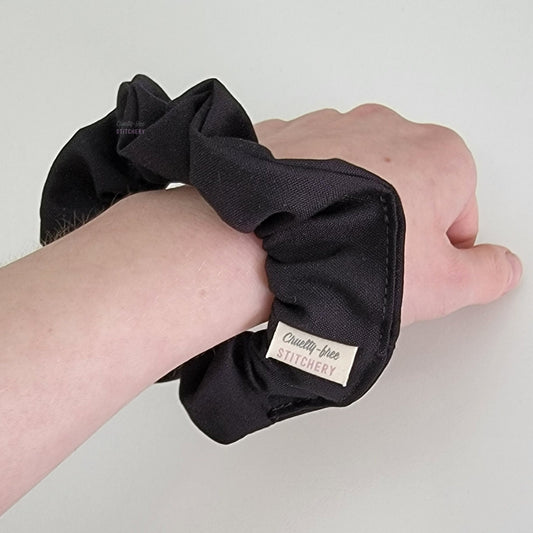 Solid black scrunchie on my wrist, with a small white tag with the Cruelty-Free Stitchery logo.