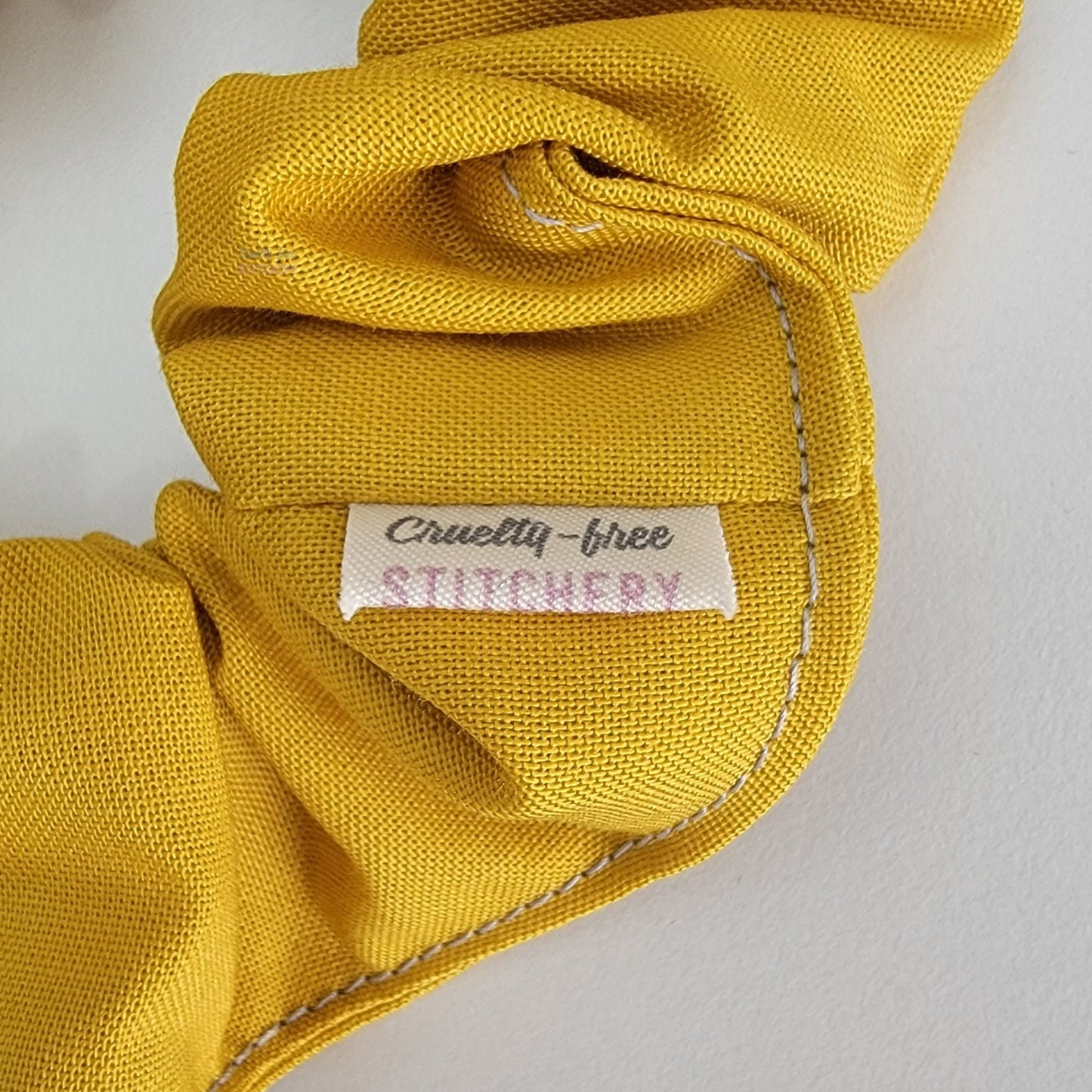 Close-up of the solid mustard yellow scrunchie with a small white tag with the Cruelty-Free Stitchery logo.