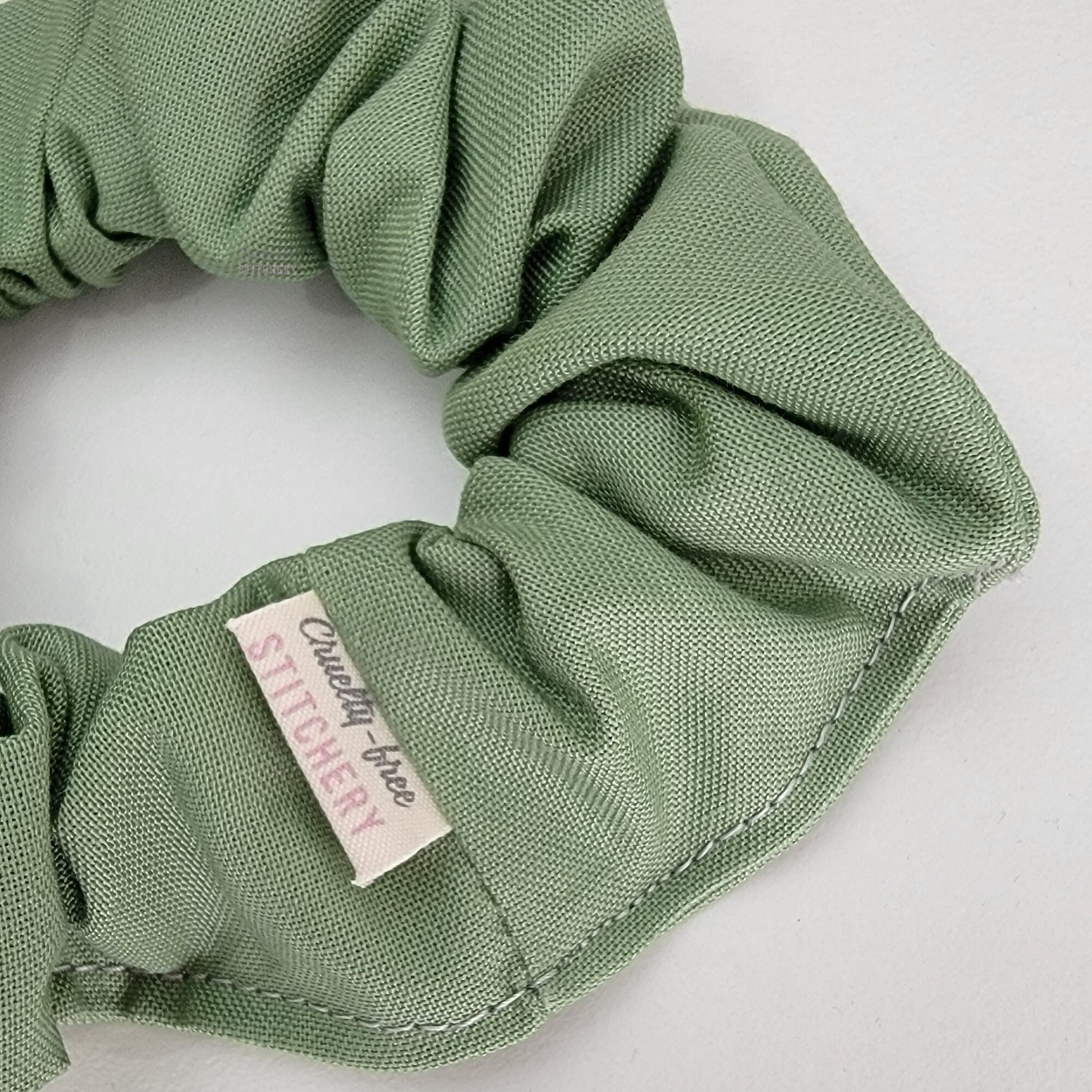 Close-up of the sage green scrunchie with a small white tag with the Cruelty-Free Stitchery logo.