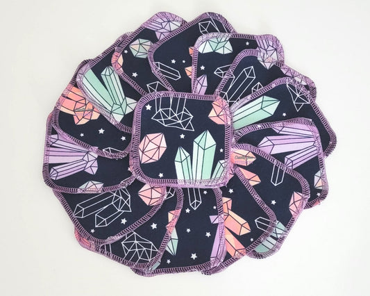 Crystals Print reusable cotton rounds, arranged in a circle. The fabric is a dark navy blue with various pink, light blue, and light purple crystals and small white stars. The rounds are a rounded square shape and stitched with light purple thread.