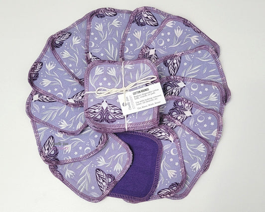 Light purple with moths print reusable cotton rounds, arranged in a circle with a bundled pack in the center. They are a rounded square shape, and stitched together with light purple thread.
