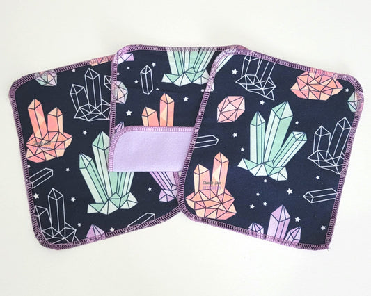 Reusable cloth wipes in a crystals print. Three wipes laid out, with one in the center folded back to show the other side is a light purple. The print on the front is a dark navy with light blue, pink, and purple crystals and tiny stars.