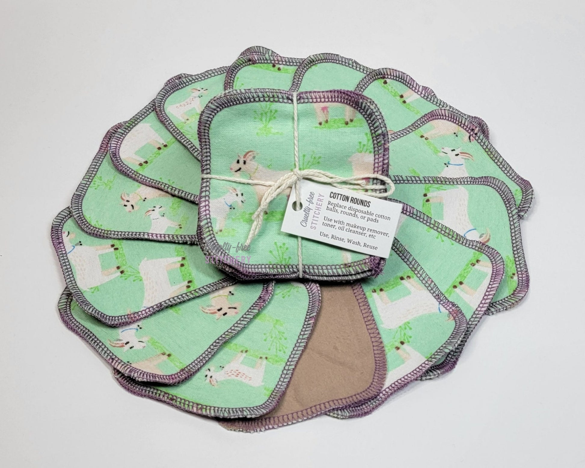 Light green with white goats print reusable cotton rounds. Arranged in a circle with a bundled pack in the center. One is flipped over to show a solid tan color on the back. They are a rounded square shape and stitched with light purple thread.