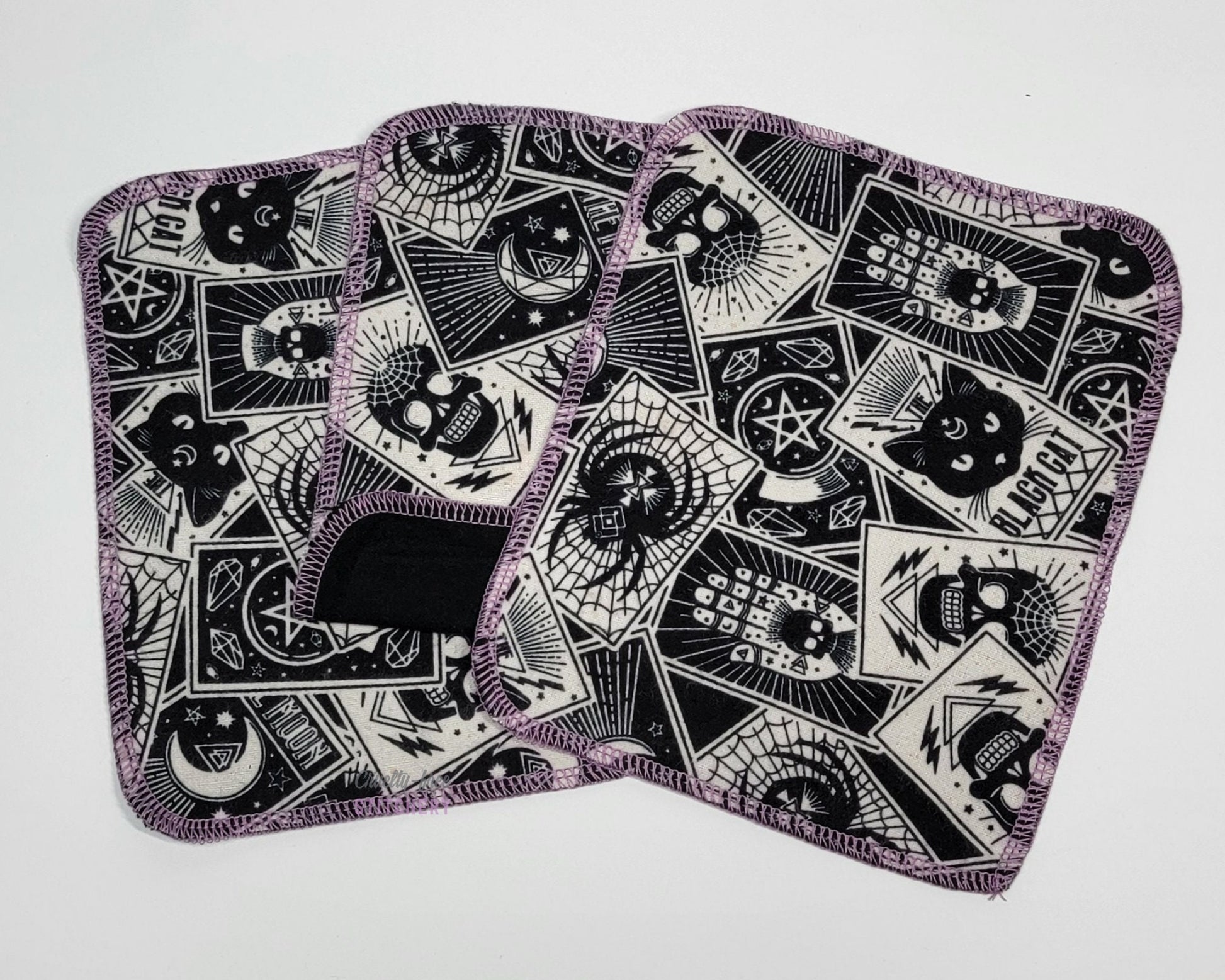 Reusable cloth wipes, a black and off-white scattered tarot cards print. Photo shows three wipes with the middle one folded back to show the back side is solid black. They are a rounded rectangle shape, stitched with lilac purple thread.