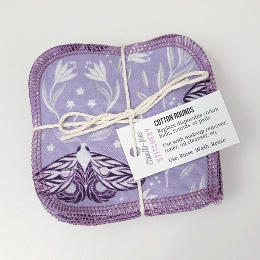 A bundled pack of moths print reusable cotton rounds, tied with a cotton string and a small paper tag. The rounds are a light purple with dark purple moths and white floral motifs.