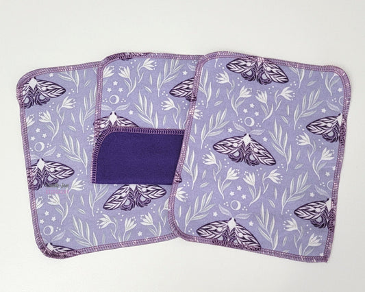 Cloth wipes - light purple fabric with dark purple moths and white floral design. Stitched with orchid purple thread. Three are arranged with one folded up to show the solid dark purple color on the back.