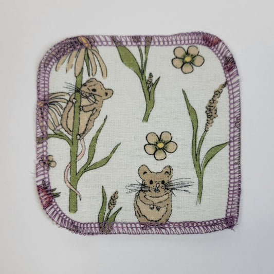 The front of one mouse print reusable cotton round. The fabric is off-white, with small light brown mice, green plants, red strawberries, and dandelions. One mouse is climbing a dandelion. 