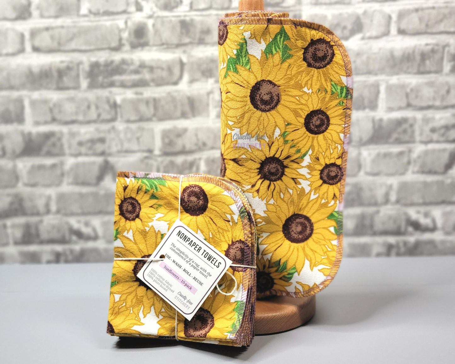 Sunflower print NonPaper Towels. Some are rolled onto a paper towel holder, next to a bundled pack. The print is bold yellow sunflowers with leaves and a white background. The edges are stitched with a light purple thread.